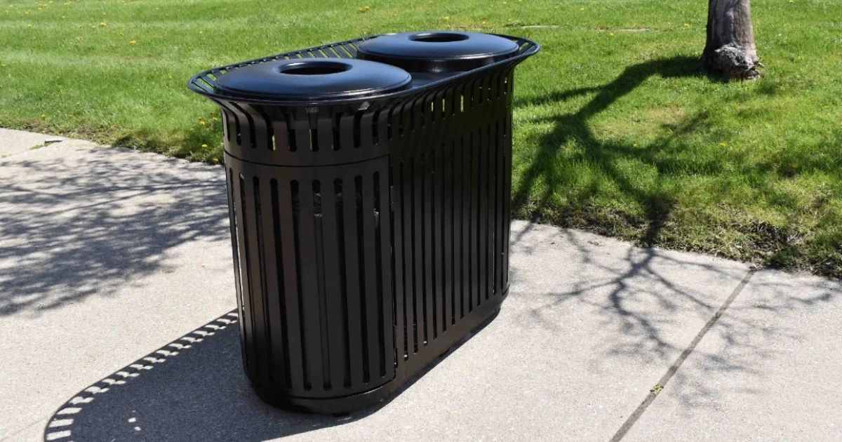 Black double garbage bin with dome lids placed on a concrete path in a grassy park area.
